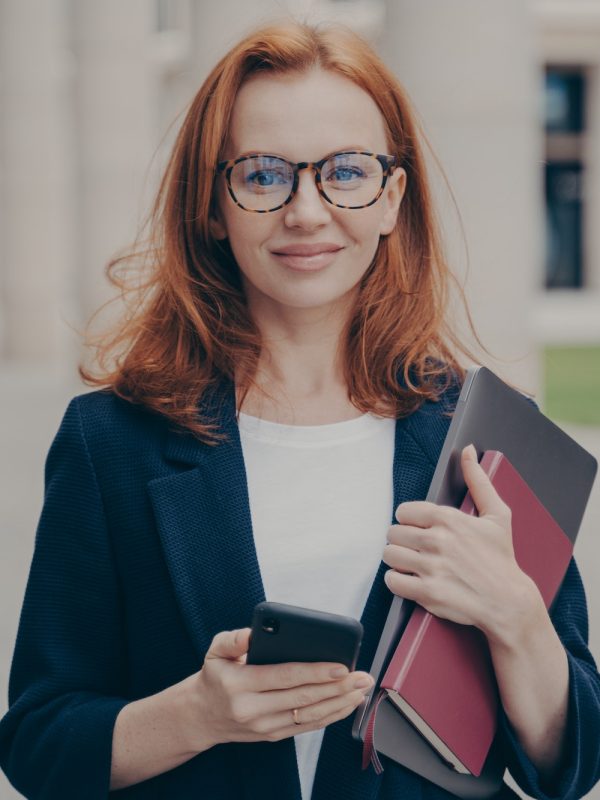 Confident beautiful red-haired female business consultant holding modern smartphone and laptop