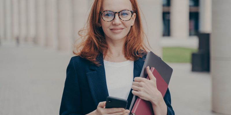 Confident beautiful red-haired female business consultant holding modern smartphone and laptop